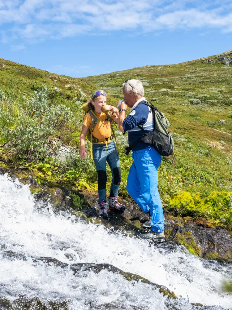Bestefar og barn tar en drikkepause med fjellvann fra bekken under fjelltur