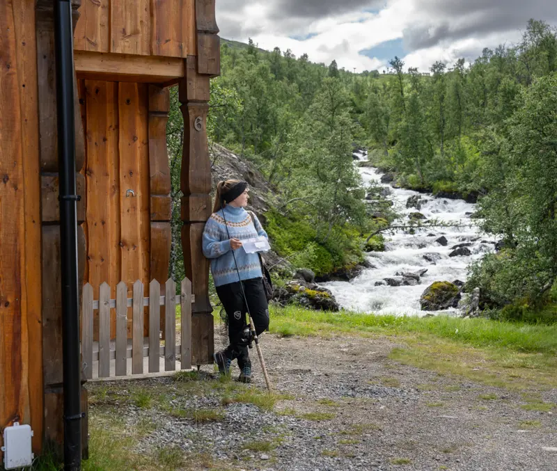 Ung kvinne med fiskeutstyr ser på elven ved siden av en av leilighetene på Filefjellstuene