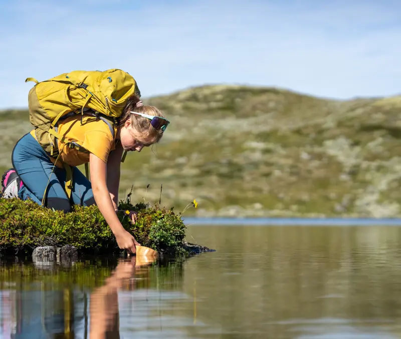 Ung jente fyller koppen med rent fjellvann under fjelltur