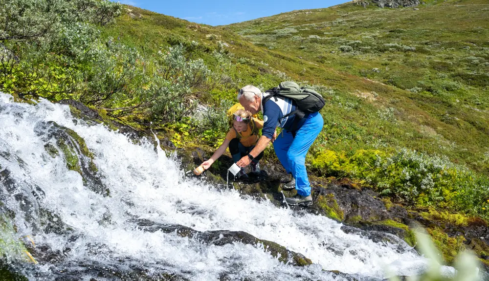 Bestefar og barn tar en drikkepause med fjellvann fra bekken under fjelltur